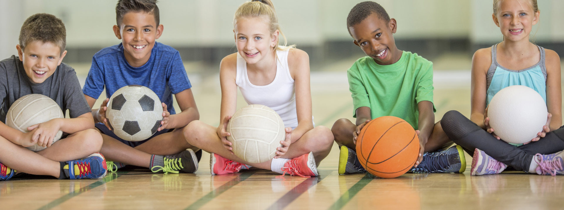 A multi-ethnic group of elementary age children are sitting in a row on the basketball court - they are smiling and looking at teh camera.